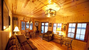 L’intérieur chaleureux d’un chalet avec un plancher en bois, un canapé, une table à manger avec des chaises et de grandes fenêtres donnant sur un lac.
