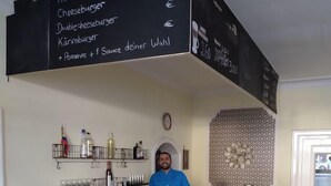 Un mostrador con un menú que enumera artículos como Hamburguesa, Cheeseburger y Doble Cheeseburger. Hay estantes con botellas y un reloj en la pared.