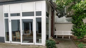 Un edificio con puerta de acceso de vidrio, un pequeño jardín con banco y una cerca blanca.