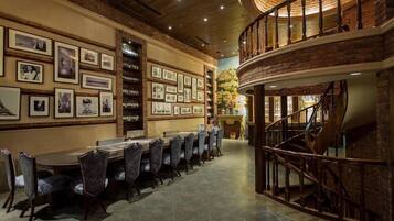 A dining area with a long table and chairs, a brick wall with framed pictures, and a spiral staircase.