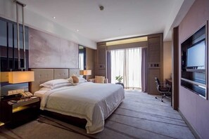 A modern hotel room with a large bed, bedside lamps, a desk with a telephone, a TV, and a sliding door leading to a balcony with a view.