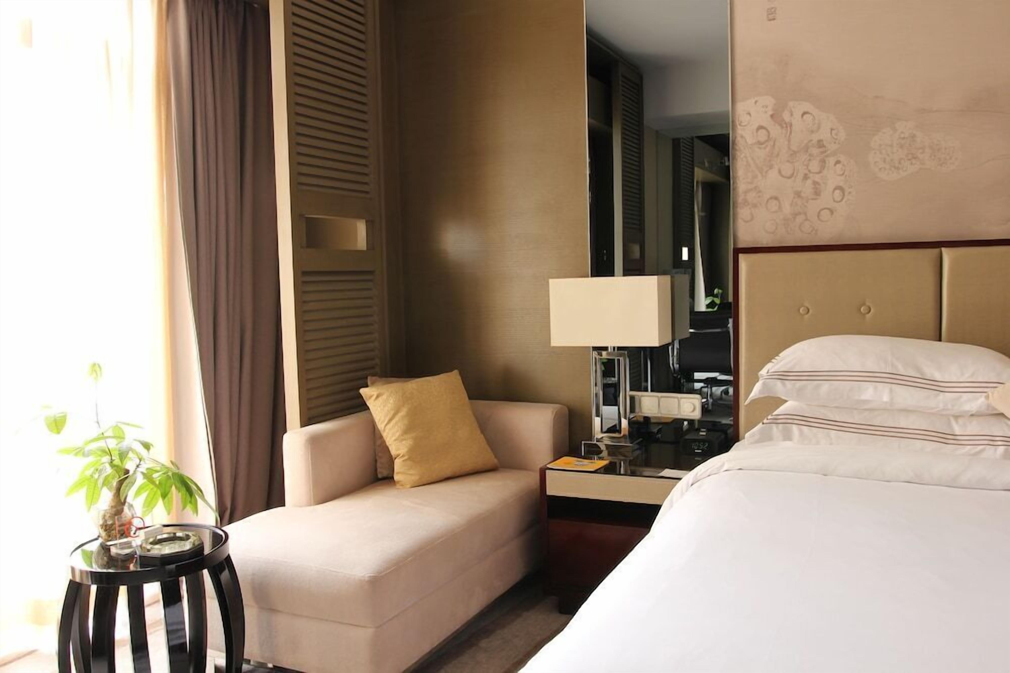 Habitación de hotel con cama, sofá, mesita de noche con lámpara, espejo y planta en maceta.