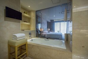 A modern bathroom with a large bathtub, a wooden stool, a towel, and a mirror reflecting a bedroom.
