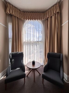 Quarto de hotel com duas poltronas, uma mesa redonda pequena e uma janela grande com cortinas.