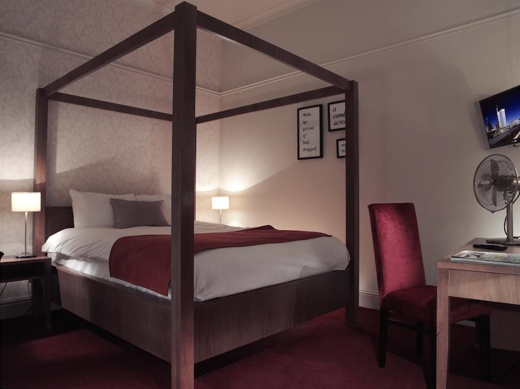 A four-poster bed with a red blanket, a desk with a chair, a wall-mounted fan, and framed pictures.