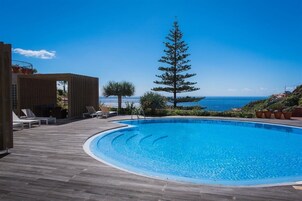 Un área de piscina con terraza de madera, sillas de descanso y vista al océano.