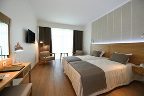 Quarto de hotel com duas camas, uma escrivaninha, uma cadeira, televisão e uma janela grande com cortinas.