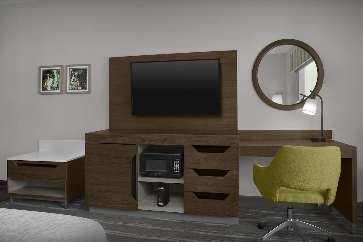 A modern bedroom with a wooden dresser, a flat-screen TV, a round mirror, and a green chair.