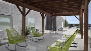 Un espace extérieur abrité, aménagé avec des chaises et des tables vertes, des poutres en bois, et offrant une vue sur un bâtiment doté de grandes fenêtres.