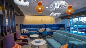 Un espace de détente moderne avec des sièges bleus, des fauteuils violets et des coussins à motifs orange. On y trouve des tables rondes et des luminaires suspendus originaux.