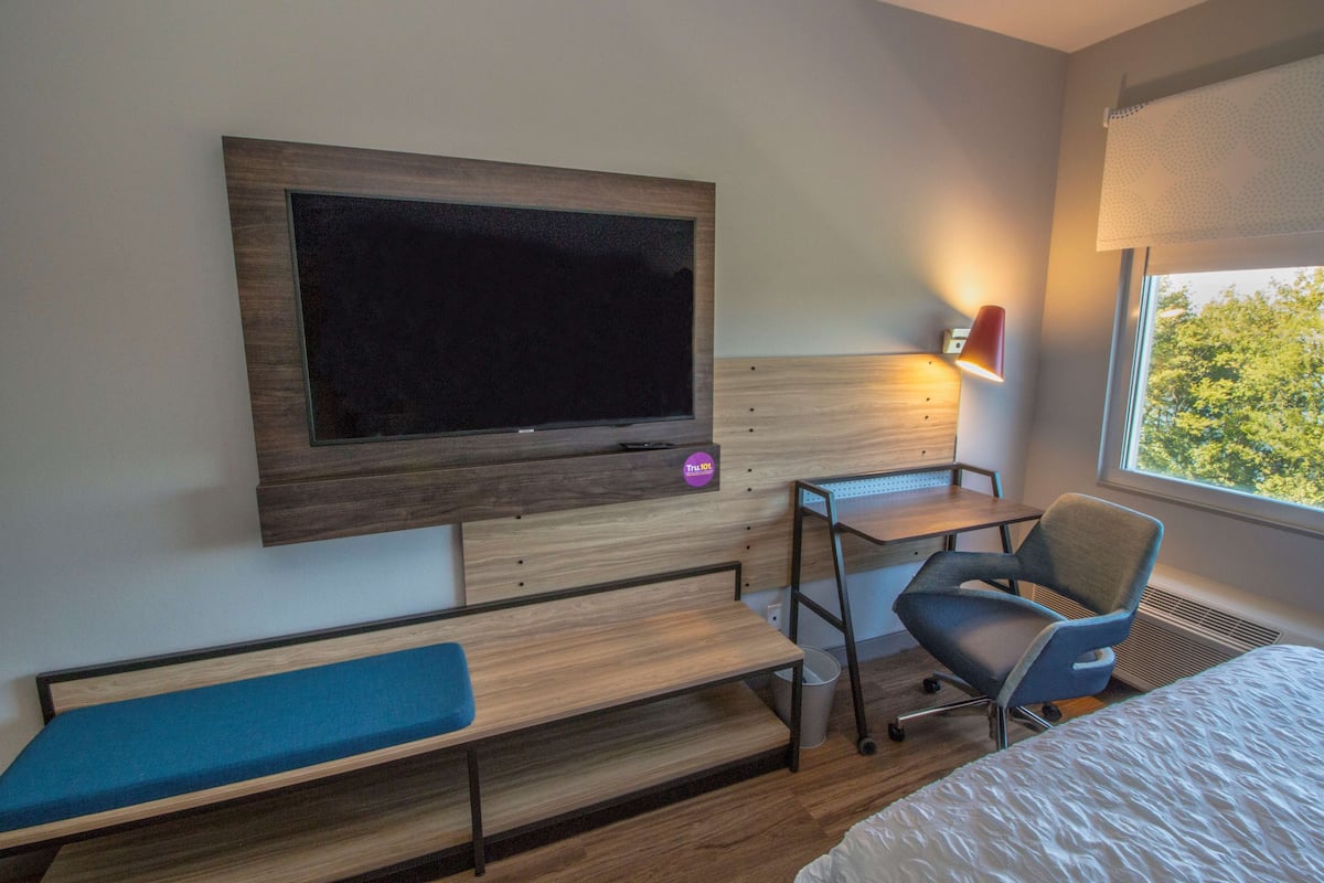 Ein Hotelzimmer mit großem Flachbildfernseher an der Wand, einem Schreibtisch mit Stuhl, einer Bank und einem Bett.