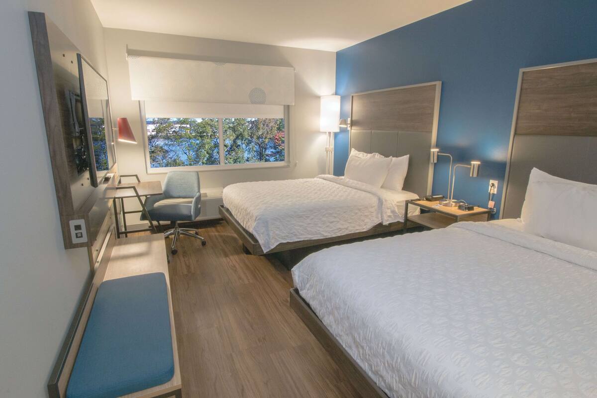 Ein Hotelzimmer mit zwei Betten, einem Schreibtisch, einem Stuhl und einem Fenster mit Blick auf Bäume.