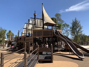 Une structure de jeux comprenant un toboggan et des éléments d’escalade, entourée d’une zone sablonneuse et de palmiers.