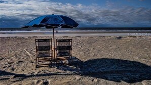 Ein Strandstuhl unter einem blauen Sonnenschirm an einem sandigen Strand, im Hintergrund das Meer.