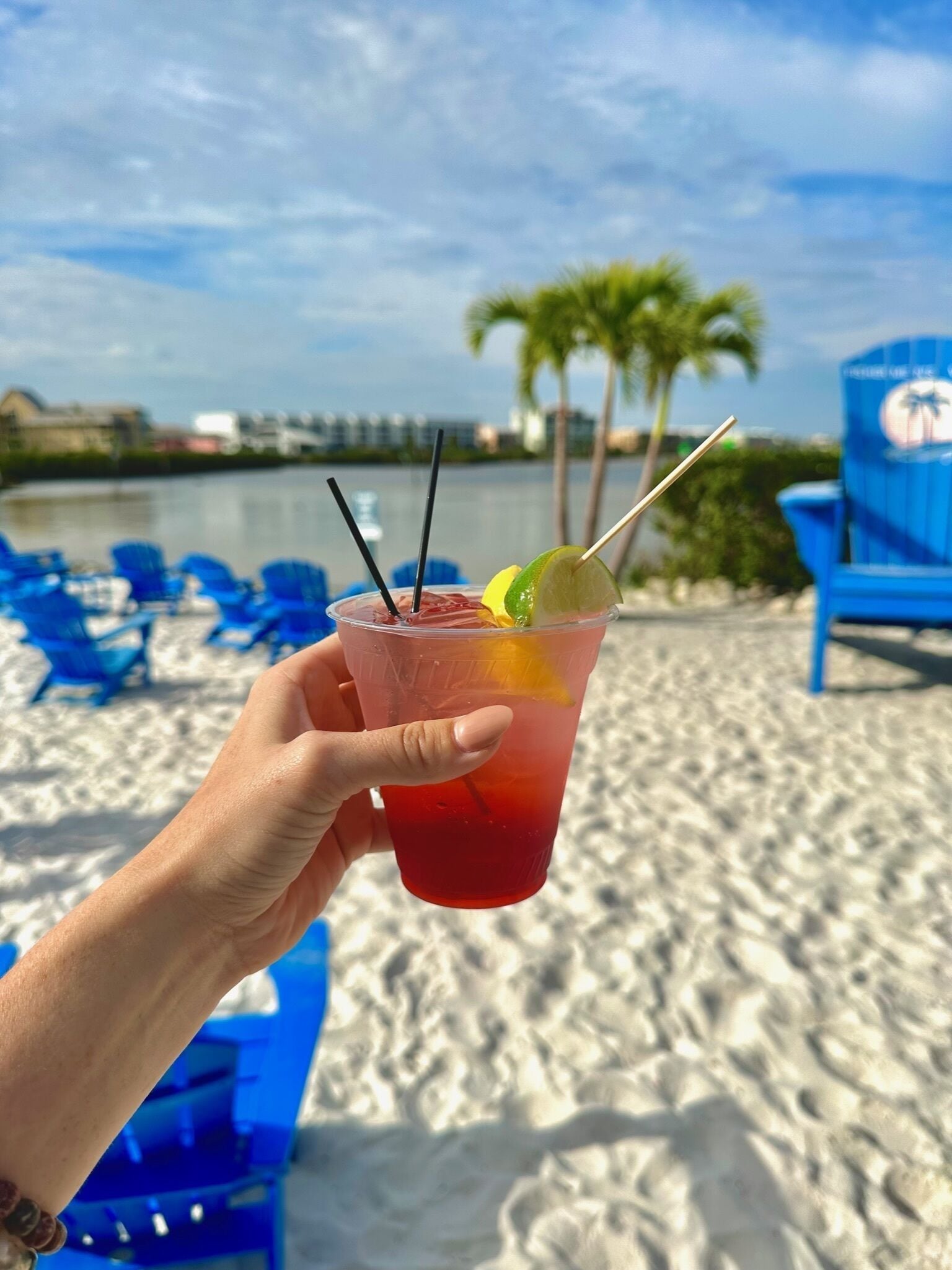Een hand vasthoudt een drankje met een limoenpartje en rietje, op het strand met palmbomen en blauwe stoelen op de achtergrond.