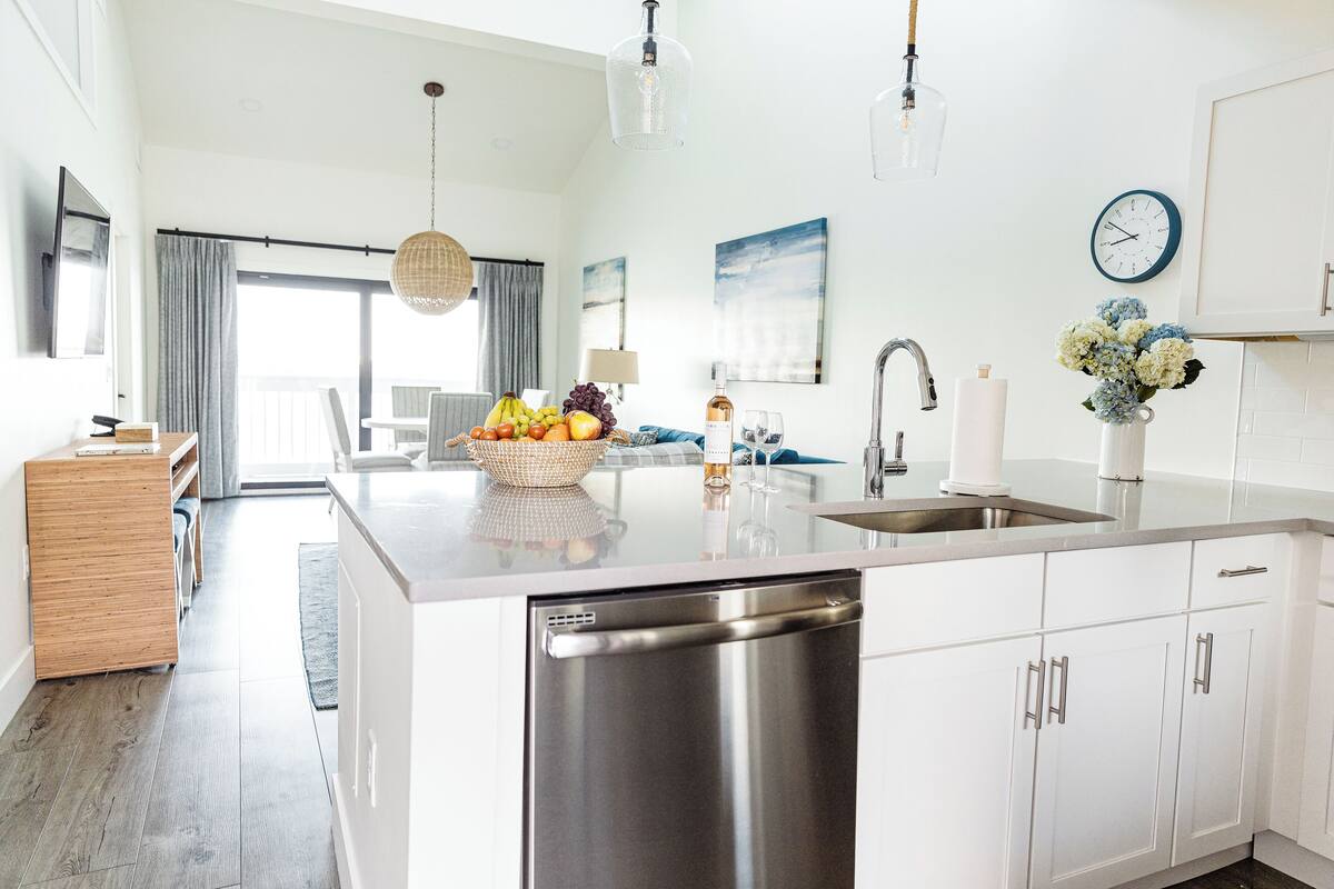 A modern kitchen with a central island, stainless steel dishwasher, white cabinetry, and a dining area with a view.
