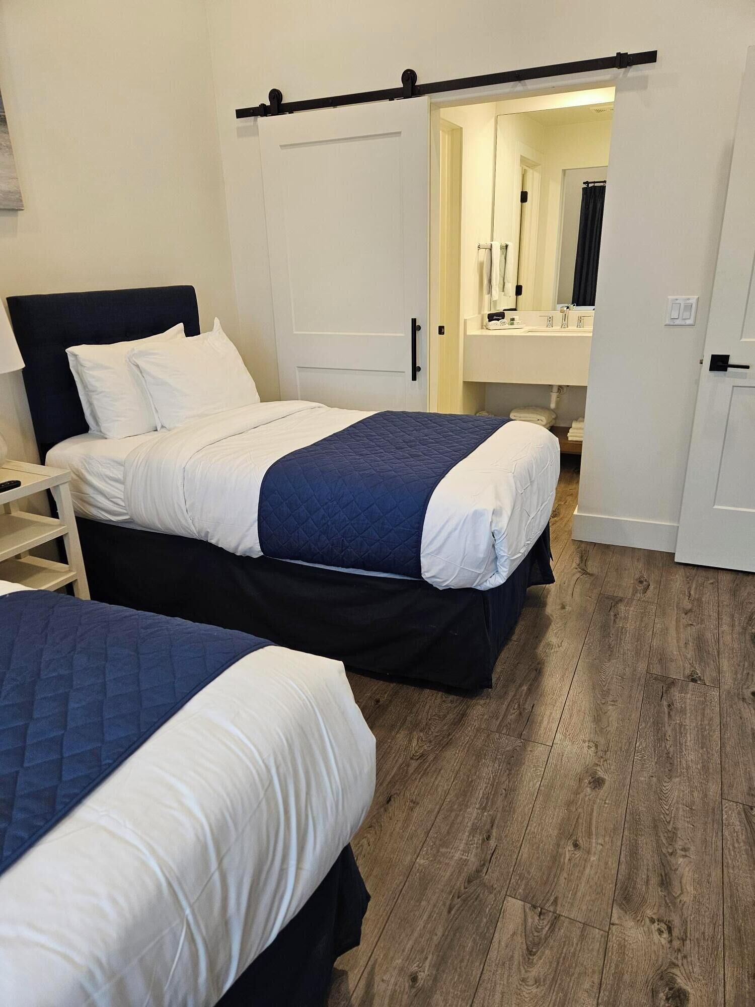 A hotel room with two beds, a wooden floor, a white door, and a bathroom visible through an open door.