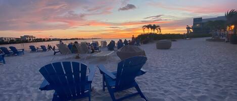 A beach with rows of blue chairs and a large umbrella, overlooking the ocean at sunset.