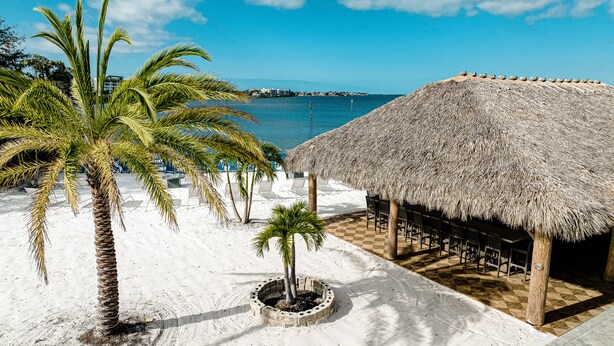 A beach with a thatched-roof structure, palm trees, and a view of the sea. - The Suites at Fishermen’s Village (Punta Gorda)