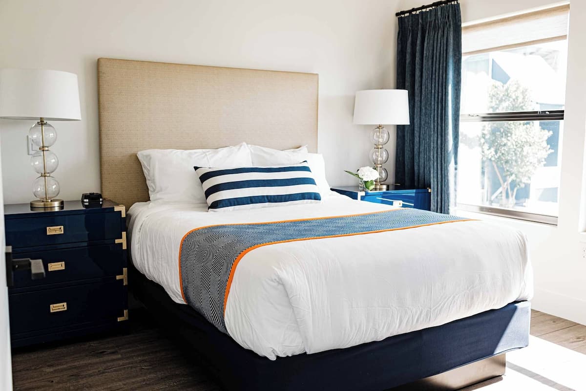 A neatly made bed with a striped pillow, a bedside table with a lamp, and a window with curtains.