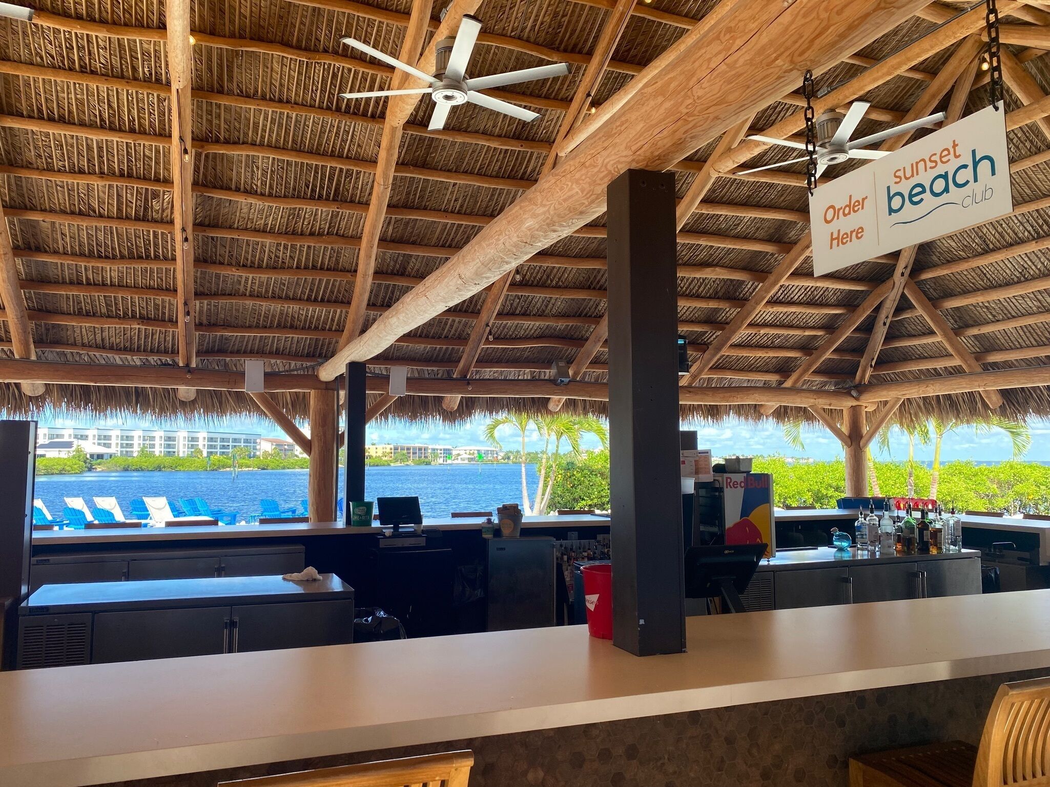 A beachside bar with a thatched roof, a sign reading "Order Here" at the bar, and a view of the ocean and palm trees in the background.