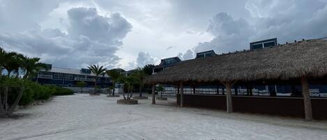 Ein Strandbereich mit strohgedeckten Hütten, Palmen und modernen Gebäuden im Hintergrund.