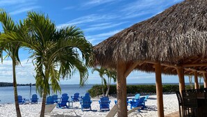 A beach with thatched-roof structures, palm trees, and blue chairs. - The Suites at Fishermen’s Village (Punta Gorda)