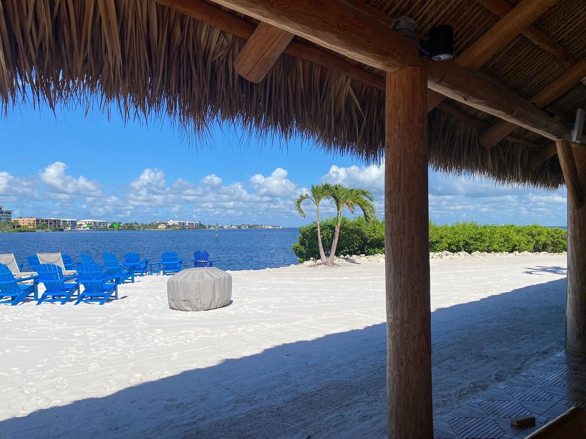 Ein Strand mit blauen Stühlen, einer überdachten Sitzgelegenheit und Blick auf das Meer und Gebäude in der Ferne.