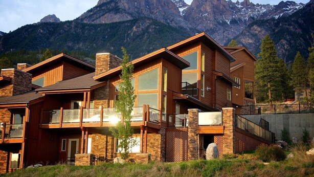 Una casa moderna de varios pisos con ventanas amplias y un balcón, con montañas al fondo.