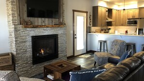 A living room with a stone fireplace, a television mounted above it, and a kitchen area with wooden cabinets and a microwave.