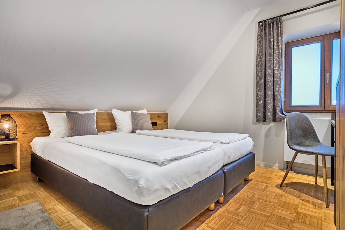 A bedroom with a large bed, a wooden headboard, a nightstand with a lamp, a window with curtains, and a chair.