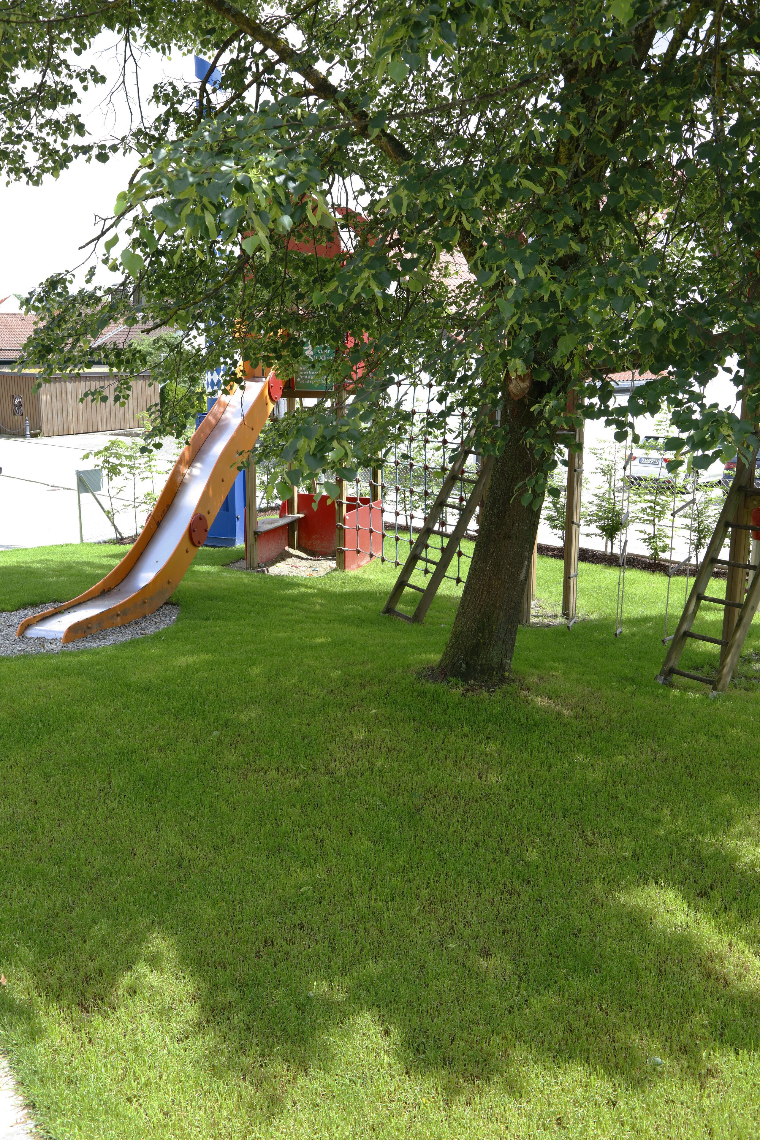 Ein Spielplatz mit Rutsche und Schaukeln, umgeben von Gras und Bäumen.