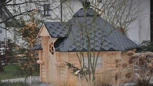 Una cabaña de madera con techo inclinado, rodeada por un jardín con diversas plantas y un pequeño árbol iluminado.
