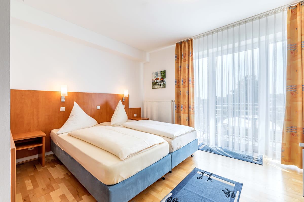A hotel room with a double bed, wooden headboard, bedside tables, a radiator, and a large window with curtains.