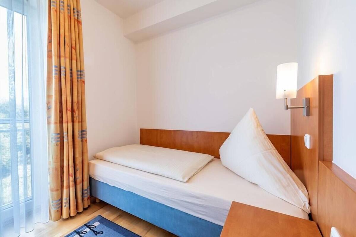 A single bed with a wooden headboard, a bedside table, a lamp, and a window with curtains.