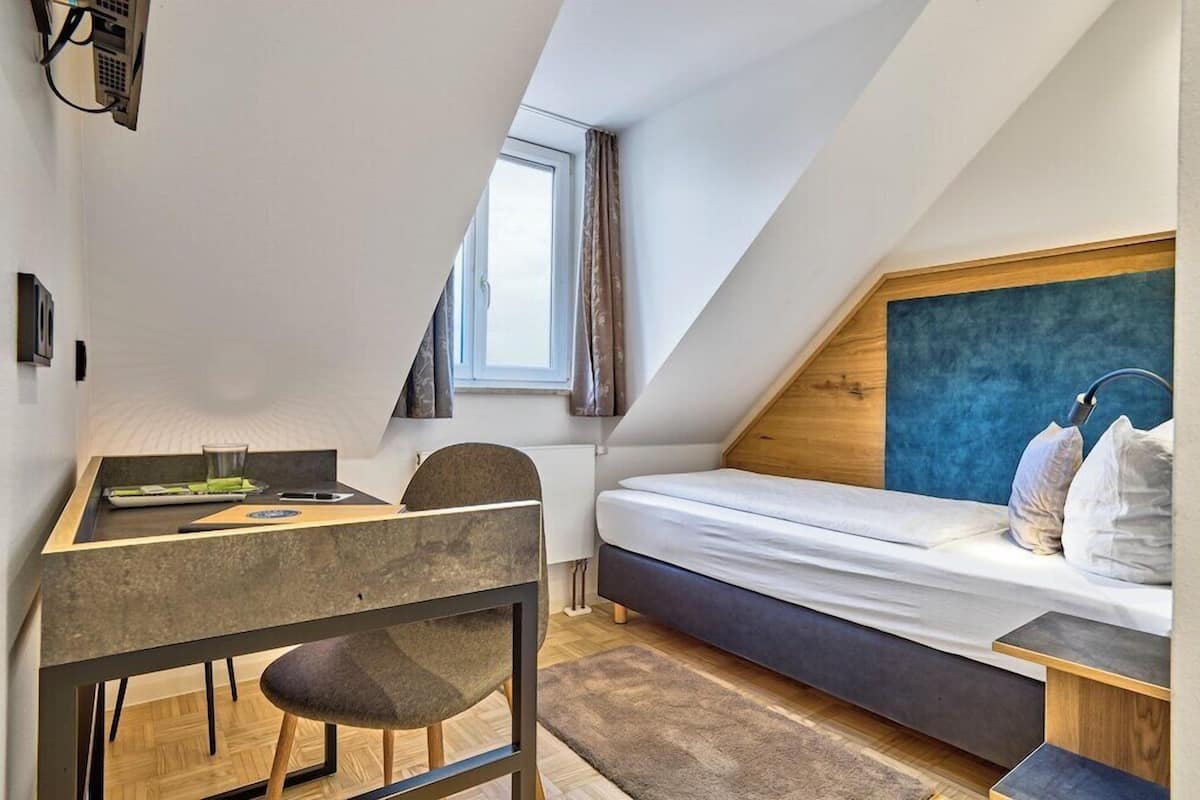 A small, modern room with a sloped ceiling, a bed, a desk, and a window.