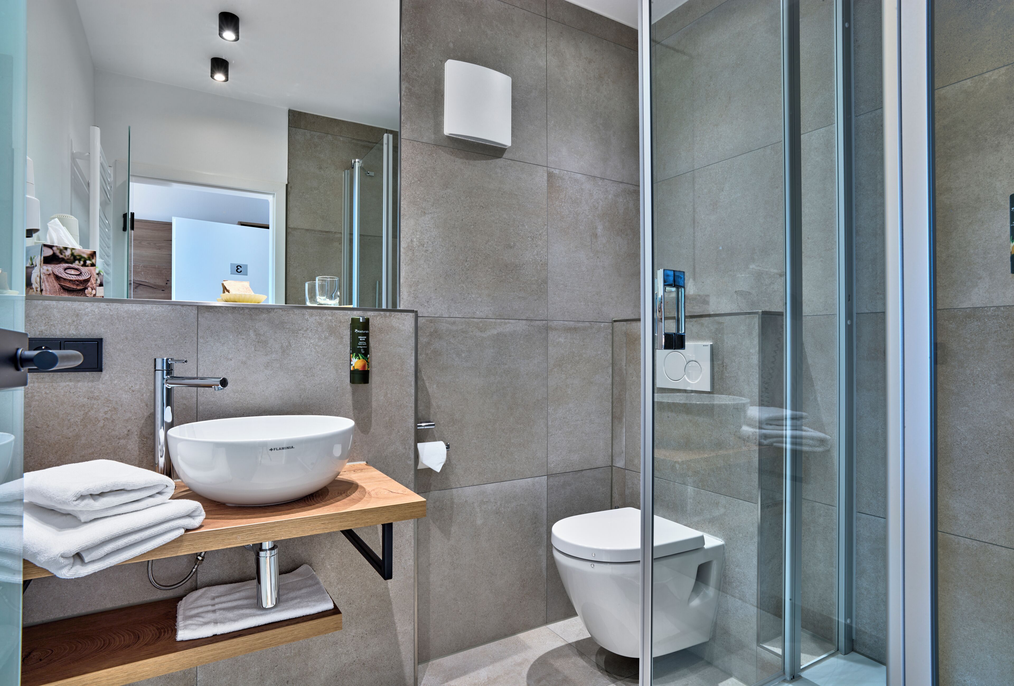 Un baño moderno con un lavamanos grande de color blanco, un inodoro y una ducha con mampara de vidrio.