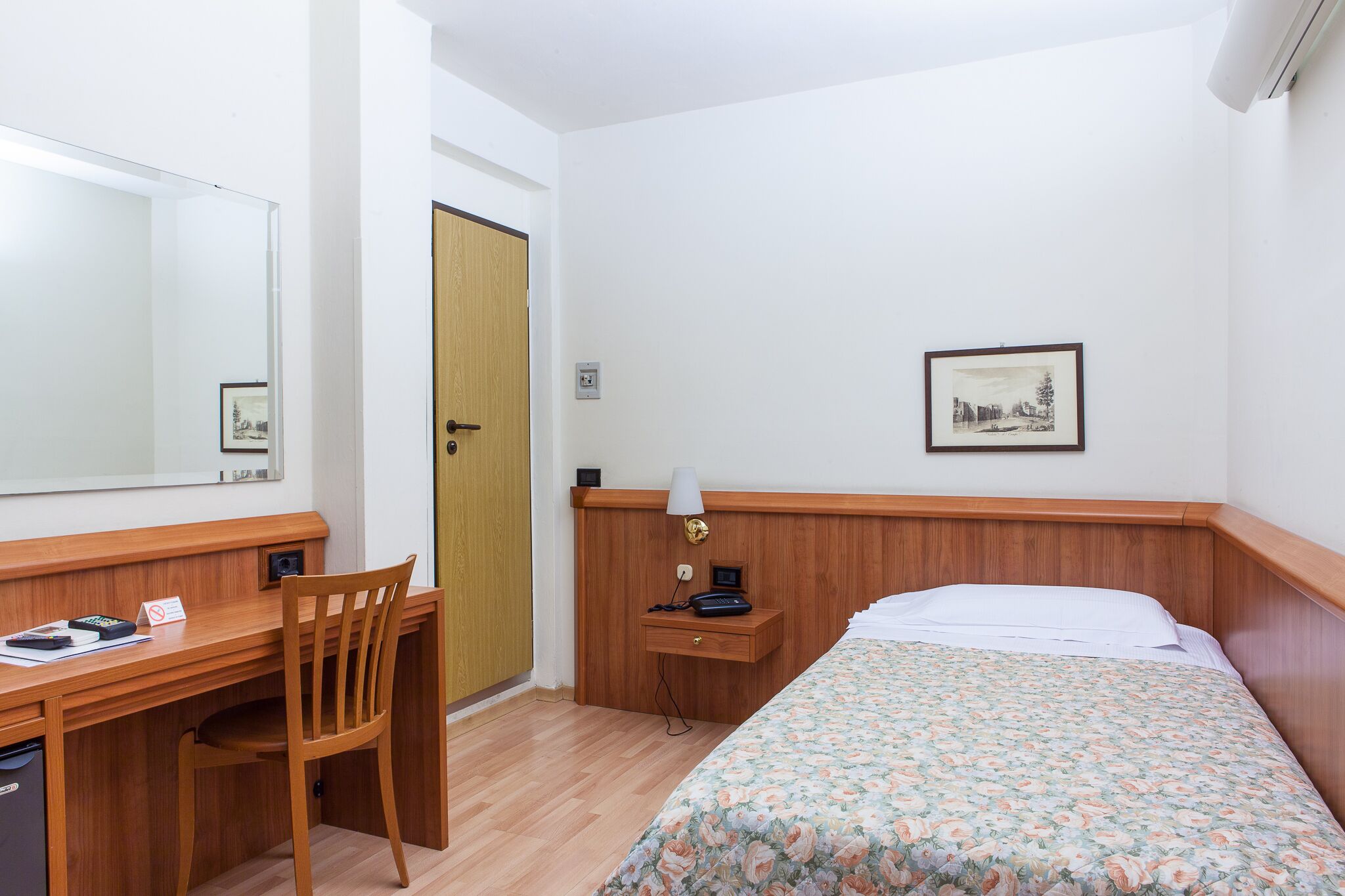 Une chambre d’hôtel avec un bureau en bois, une chaise, un lit recouvert d’une courtepointe à motifs floraux, une table de chevet avec une lampe et un tableau encadré accroché au mur.