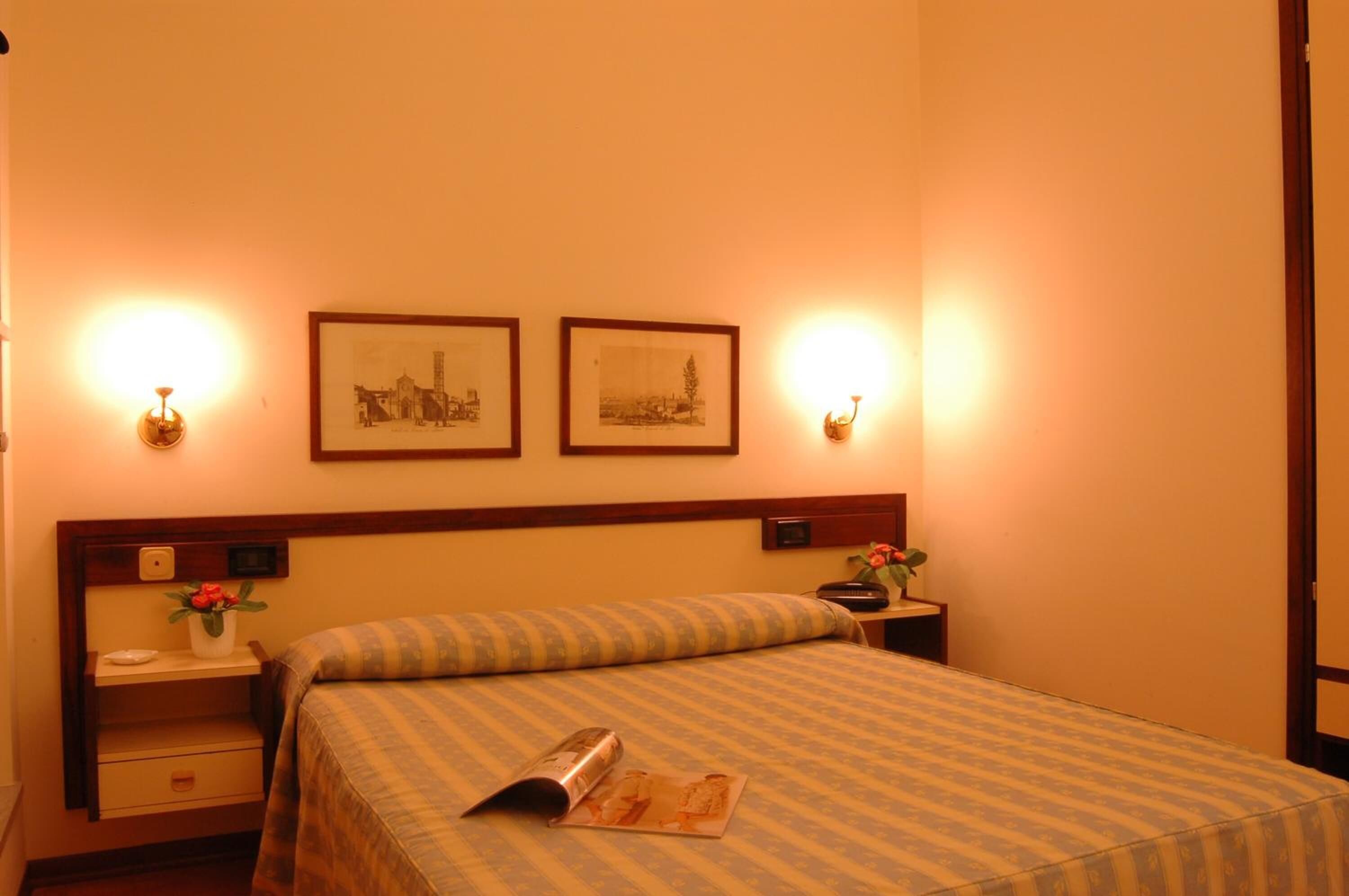 A hotel room with a bed, bedside tables, framed pictures on the wall, and wall-mounted lamps.