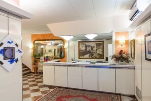Un espace d’accueil avec un sol à carreaux, un comptoir d’accueil, un miroir, des tableaux encadrés et une plante en pot.