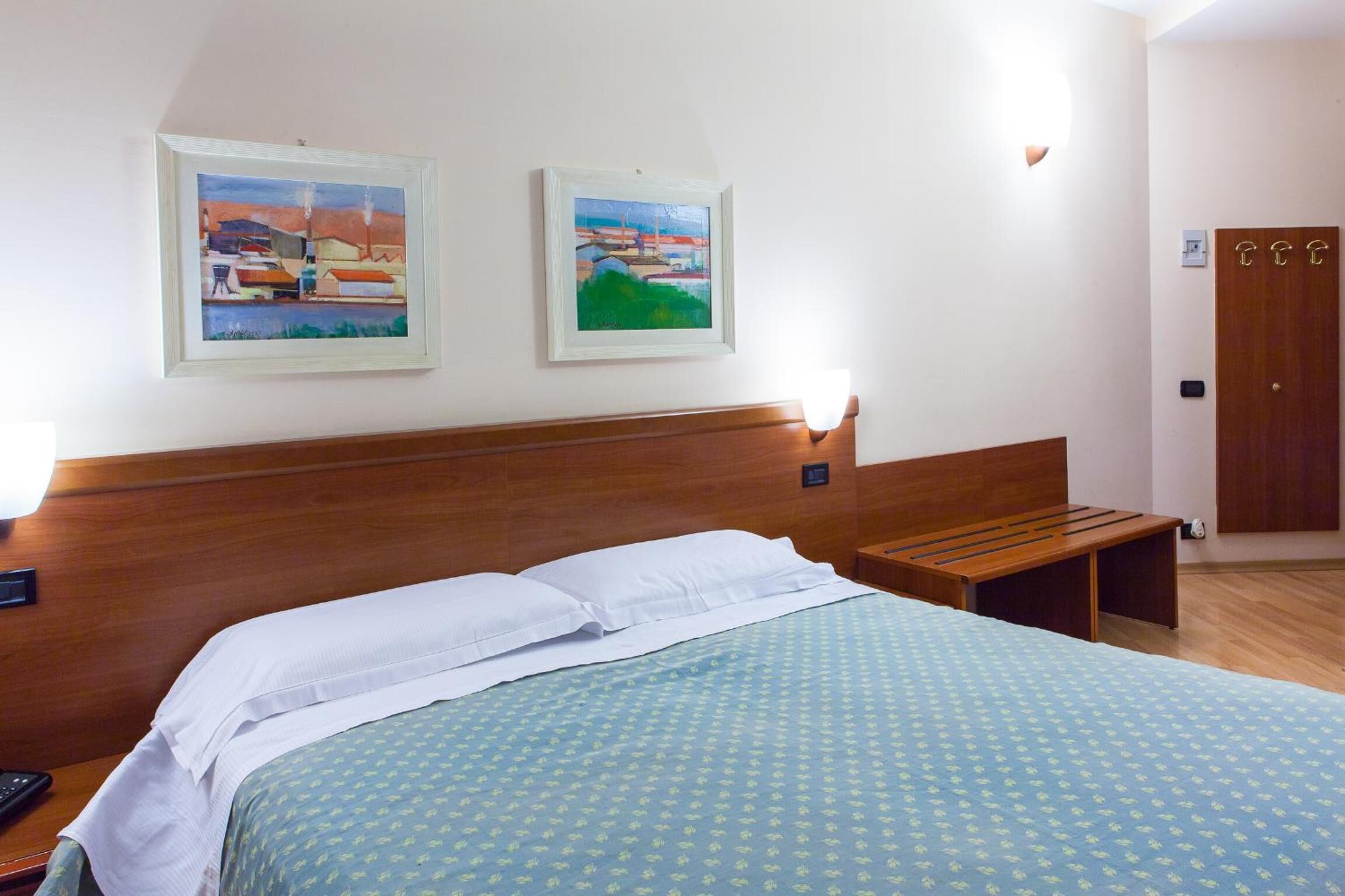 A hotel room with a bed, two framed paintings on the wall, a wooden bench, and a door.