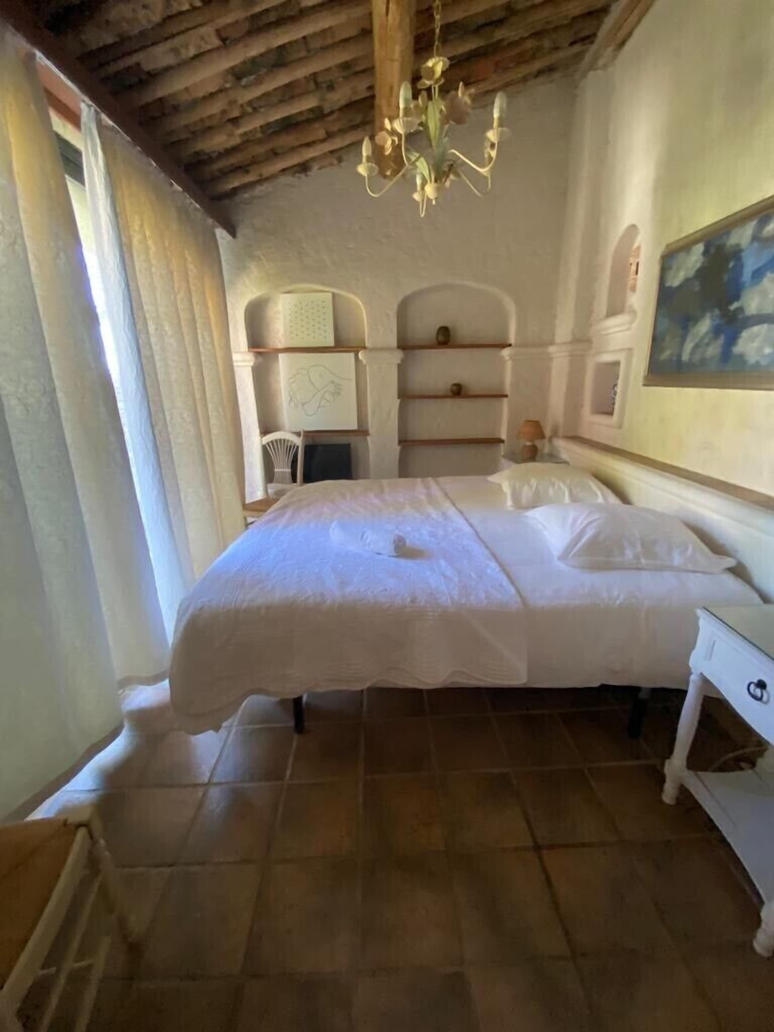 A bedroom with a large bed, a chandelier, a painting on the wall, and a window with curtains.