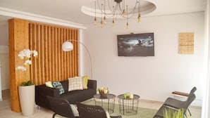 A modern living room with a black sofa, a wooden accent wall, a large potted plant, a round coffee table, and a TV mounted on the wall. - Ouril Hotel Julia (Sal)