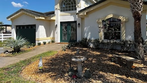 Kaksikerroksinen talo, jossa on vihreä ovi, autotalli ja kauniisti maisemoitu etupiha, jossa on lintulautaa ja palmu.