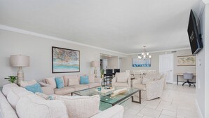 A modern living room with a glass coffee table, beige sofas, and a dining area with a chandelier. - Oceanfront Corner - Horizons Condo #1701 (Daytona Beach)
