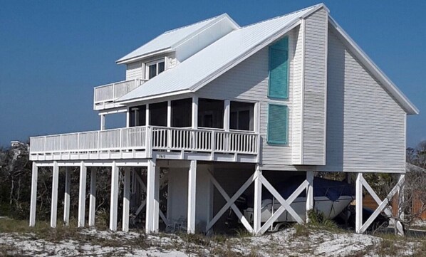 A two-story house with a balcony and a covered area, built on stilts over a sandy area.