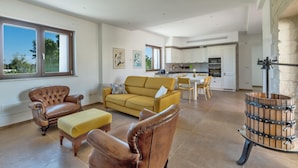 Een ruime woonkamer met een bank, een leren fauteuil en een voetenbankje. In de achtergrond is een keuken te zien met witte kastjes en een eethoek.