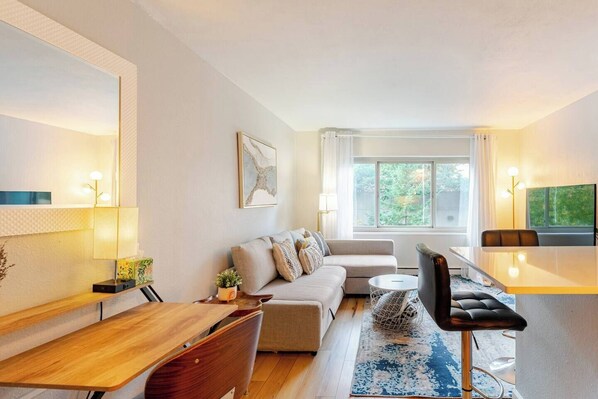 A modern living room with a sofa, coffee table, and dining area. There is a large window providing natural light and a view of greenery outside. - Serene downtown Seattle condo - Walk to Convention Center, Pike Place, top sites (Seattle)