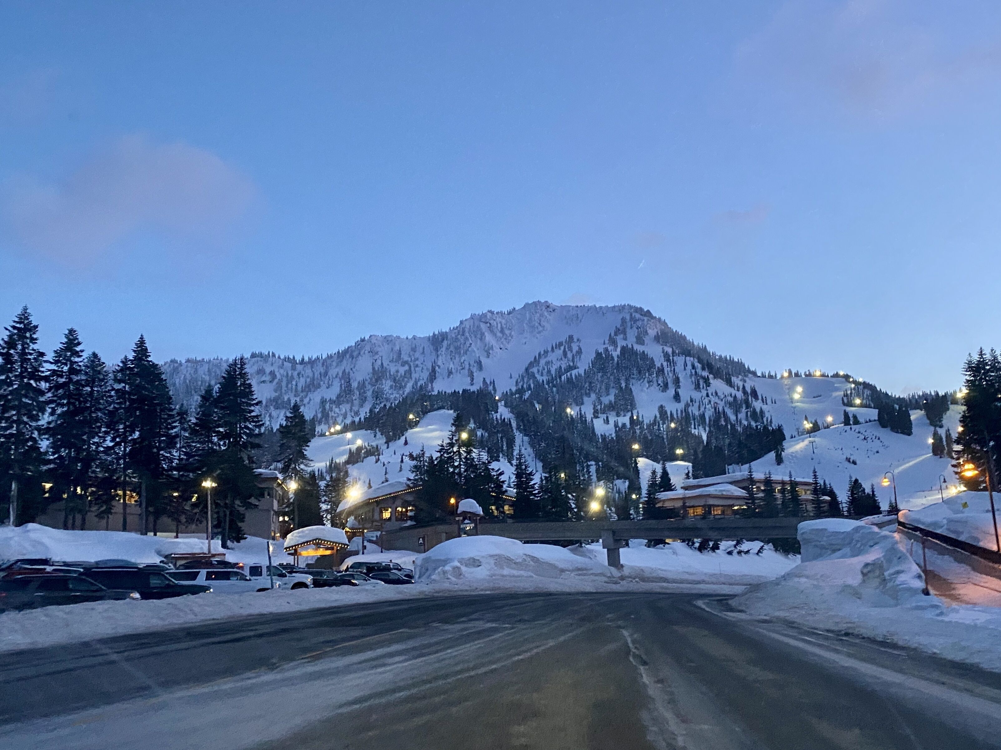 Eine schneebedeckte Straße mit einem Berg im Hintergrund, beleuchtete Straßenlaternen und geparkte Autos.