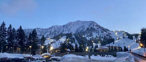 A snow-covered road with a mountain in the background, illuminated street lights, and parked cars.